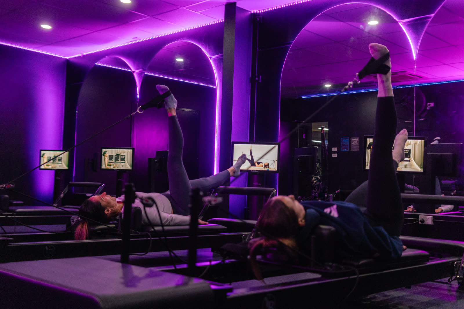 Members using reformer pilates beds under purple LED arches